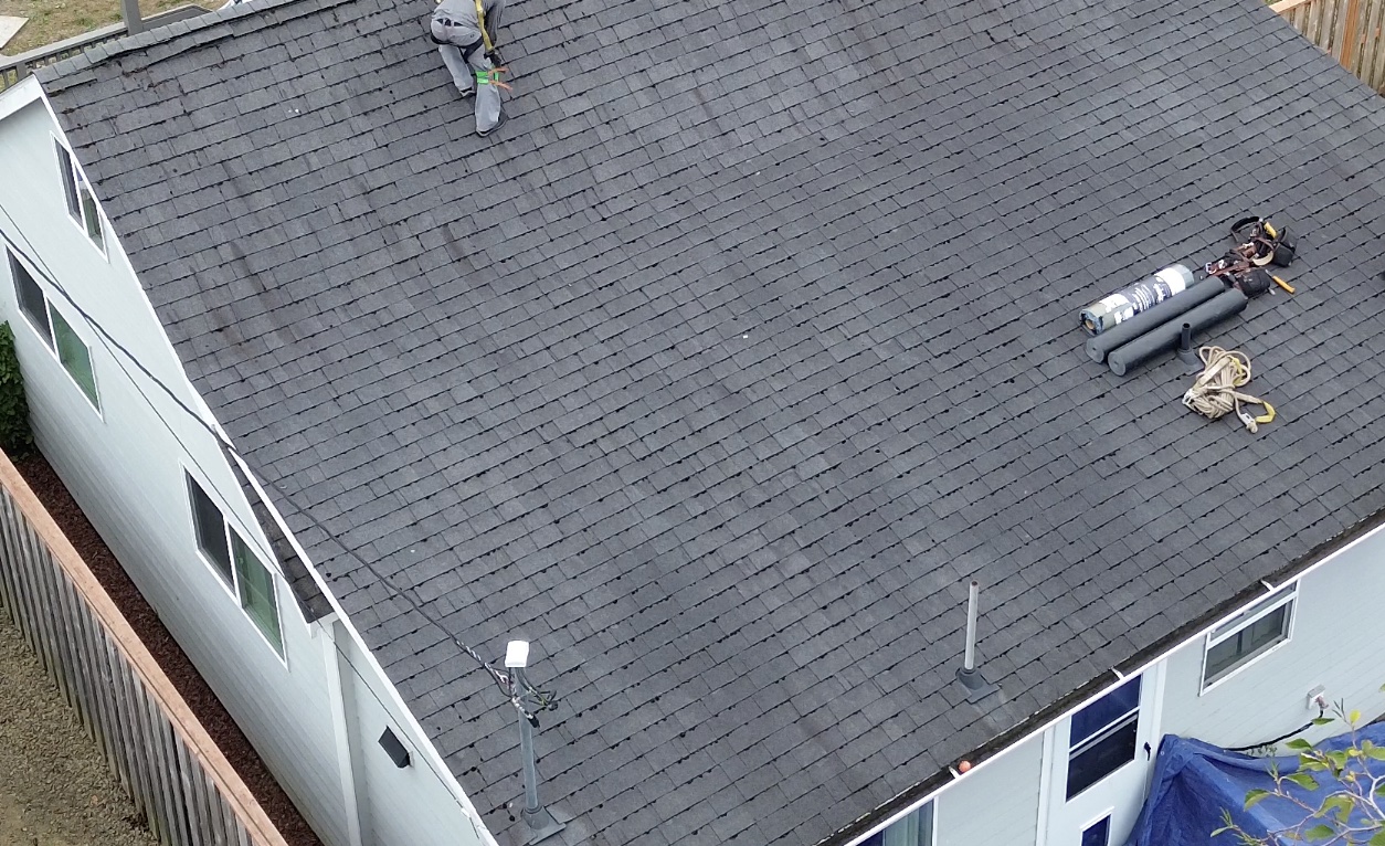 Drone view of a worn gray 3-tab roof with a crew member starting tear-off and materials staged on the slope